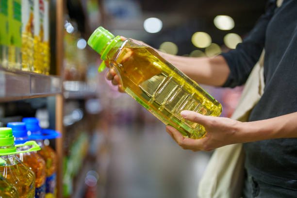 In a close-up shot, a woman's hands are reading the label on a bottle of cooking oil taken from the shelf in a grocery store, ensuring product freshness and safety.
