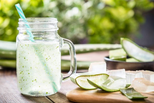 Front view of a drinking glass full of an aloe vera drink surrounded by some aloe vera crystals and sliced leaves. A black bowl full of sliced aloe vera leaves is defocused on the background. Predominant color are green and brown. Studio shot taken with Canon EOS 6D Mark II and Canon EF 24-105 mm f/4L